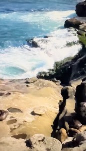 Waves crashing against rocky coastal cliffs with smooth stones and clear blue water.